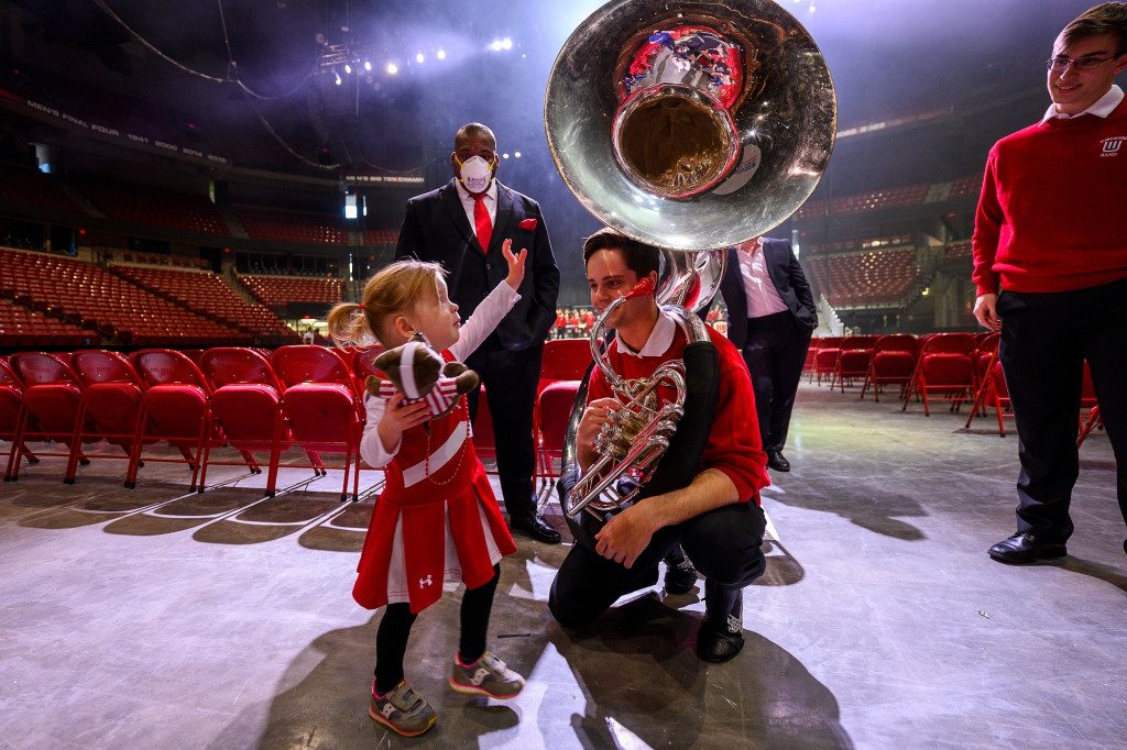 A toddler wearing a UW cheer uniform stands in a near empty Kohl Center. She holds a stuffed Bucky Badger in one hand and reaches up with her other to touch the bell of a marching band member’s sousaphone.