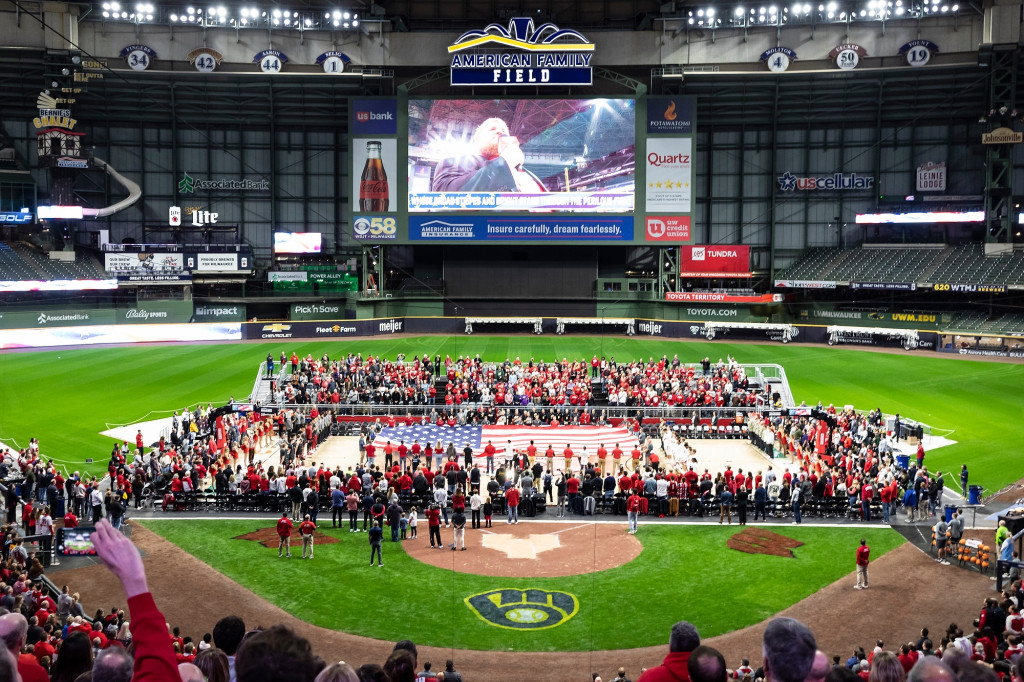 A basketball court set up in the infield at American Family Field is filled with people displaying a nearly court-sized American flag, surrounded by bleachers filled with spectators.