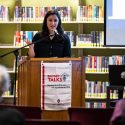 Lori Kido Lopez speaks from apodium in a library setting. A Badger Talks banner hangs from the front of the podium.