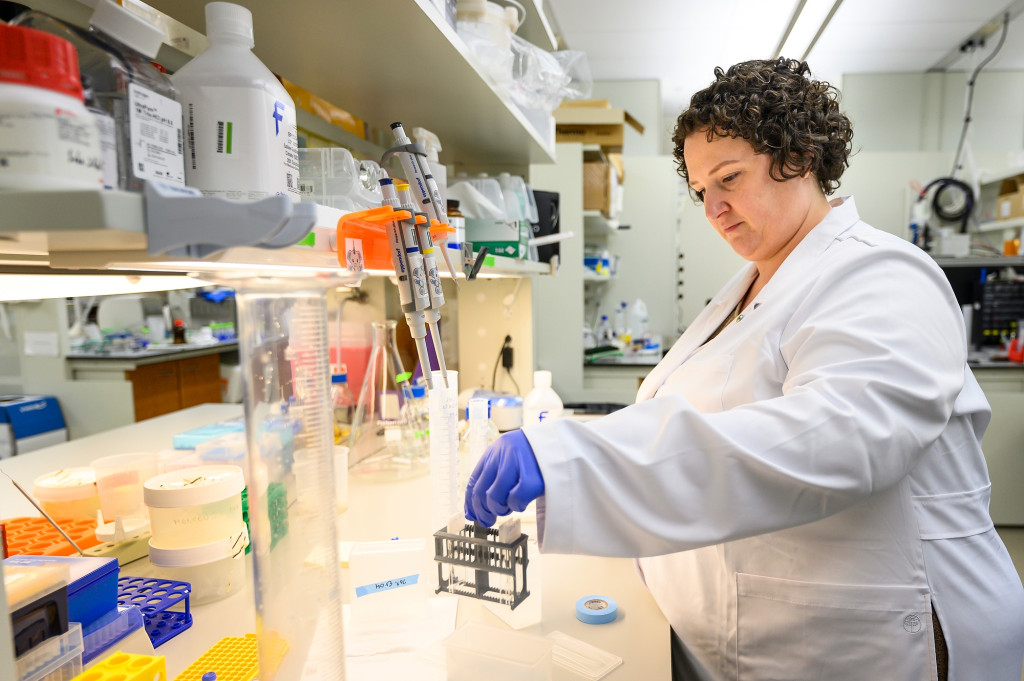 Kirstan Gimse wears a white lab coat and gloves while working with samples at a lab bench.