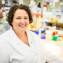 Portrait of Kirstan Gimse in a white lab coat in front of a lab bench filled with colorful equipment.