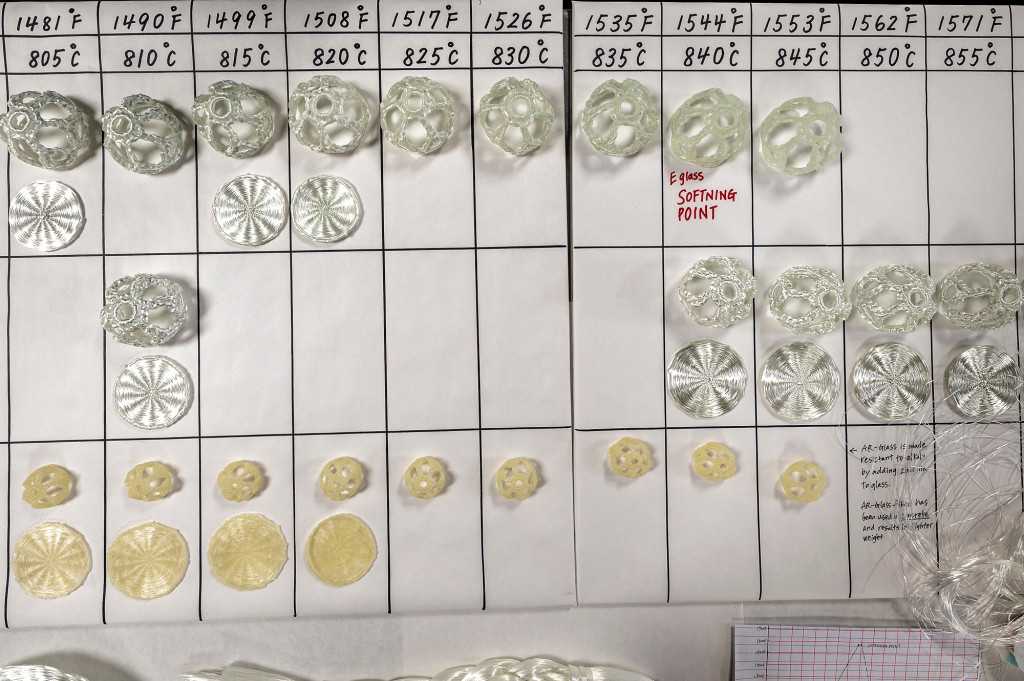 Aerial view of a table displaying finished fiberglass pieces made from different varieties of fiberglass and fired at different temperatures. Materials and temperatures are labeled, and the pieces are arranged in a grid pattern.