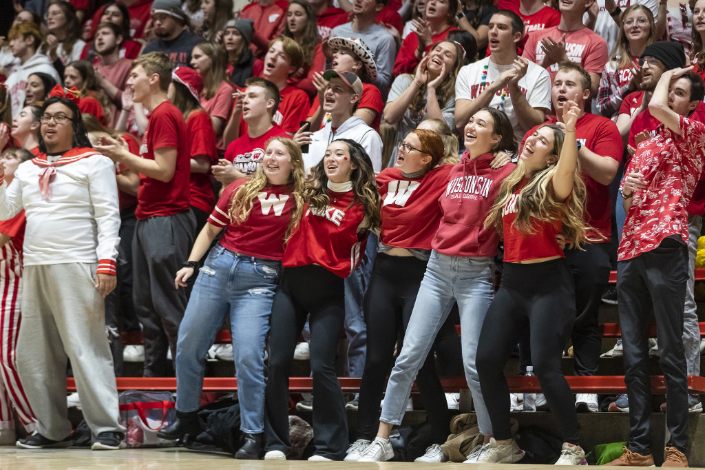 Young people wearing red Wisconsin sweatshirts stand and cheer in the stands.