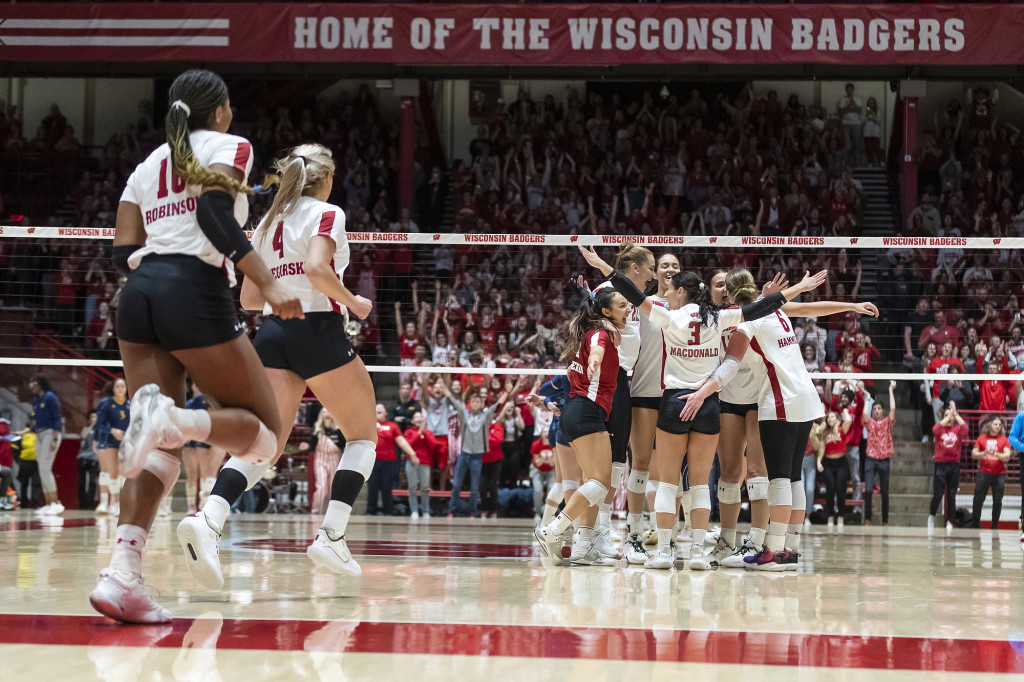 Uniformed volleyball players run jubilantly onto a court and hug, as fans cheer.