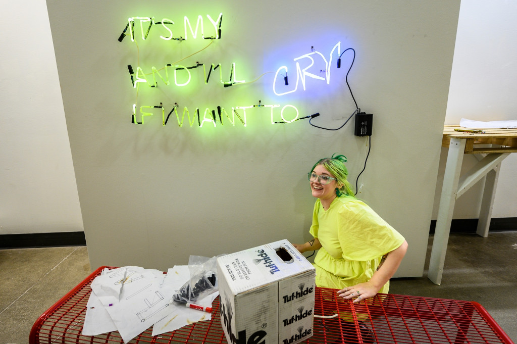 A woman smiles as she looks at a sculpture made of green and purple neon tubes.