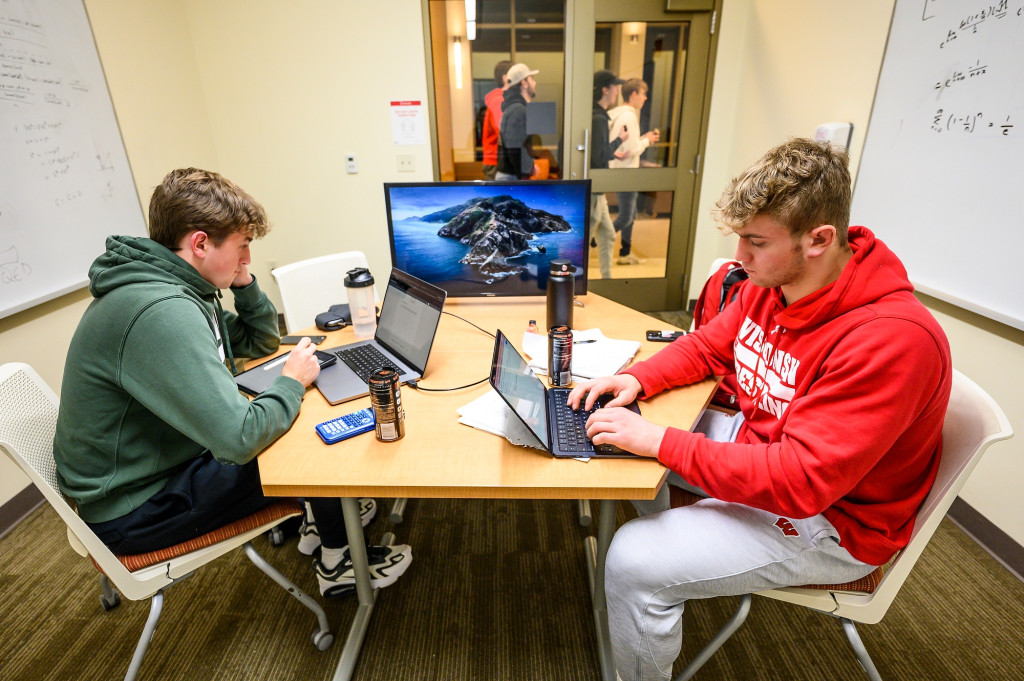 Two men sit at a table and type in the laptops.