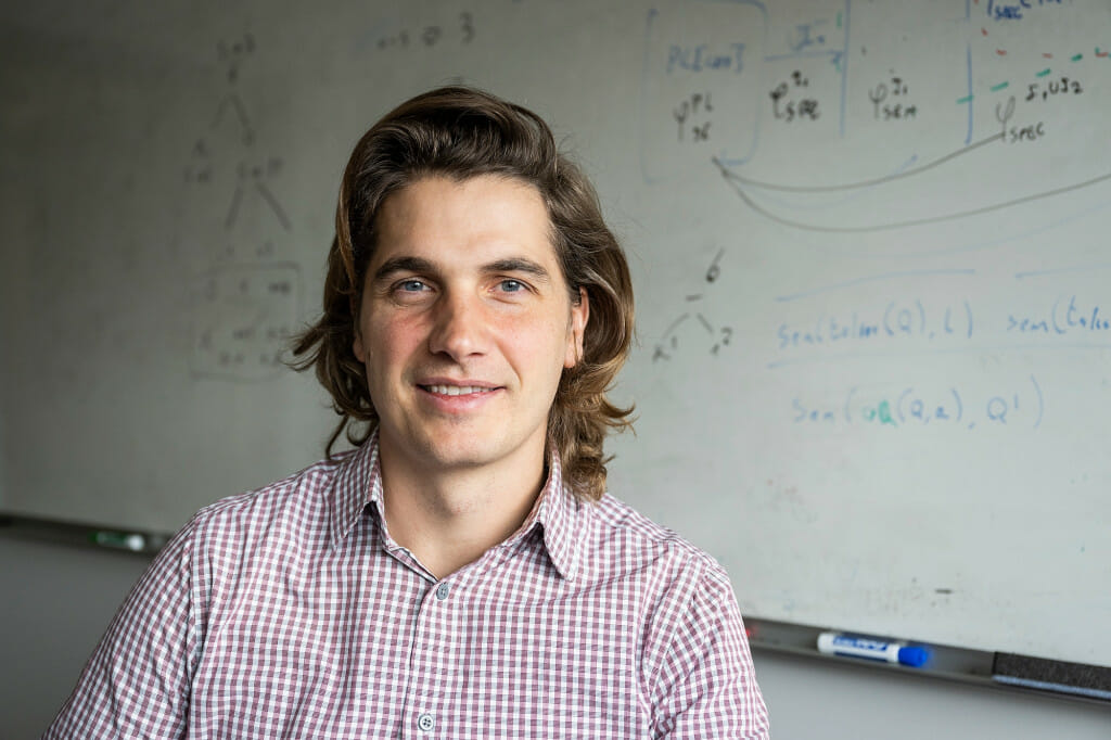 A man smiles, seated with a white board in the background.