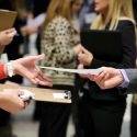 A student shares a resume with a potential employer. The photo is a closeup on the student's hand extending a resume for an employer to accept.