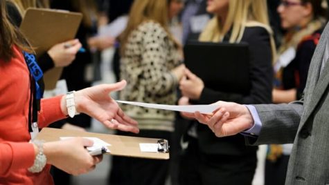 A student shares a resume with a potential employer. The photo is a closeup on the student's hand extending a resume for an employer to accept.