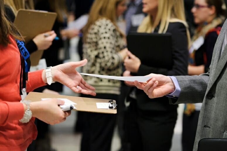 A student shares a resume with a potential employer. The photo is a closeup on the student's hand extending a resume for an employer to accept.