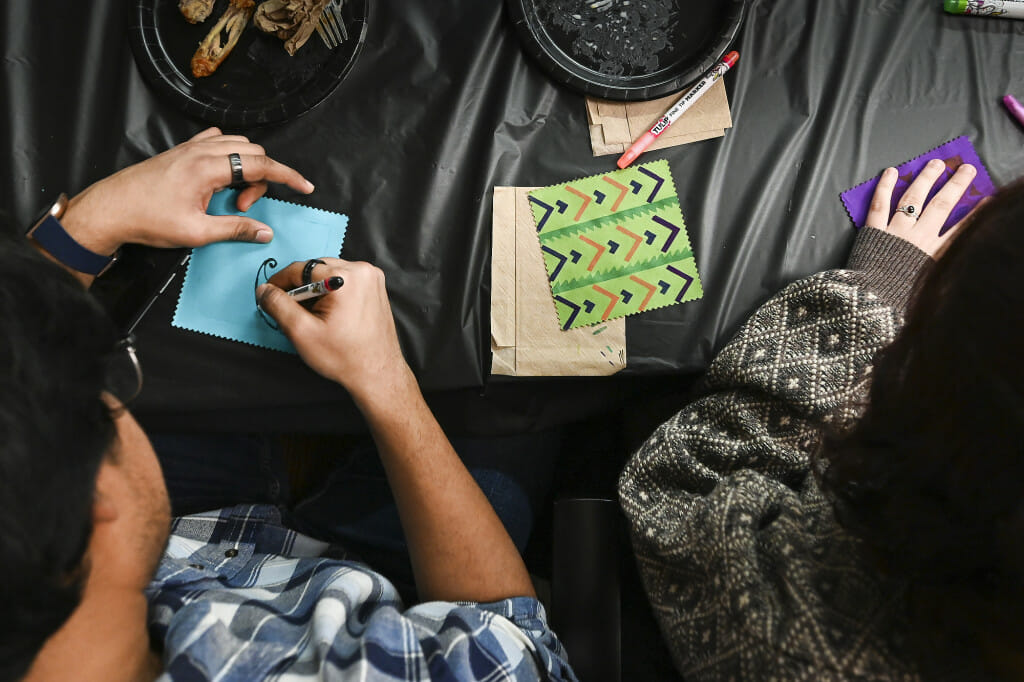 An overhead view of a table covered with a black cloth. From the lower left corner, a person leans in to draw with a marker on a blue cloth square. To their right lie a green fabric square fully decorated in black and pink along with crafting supplies.