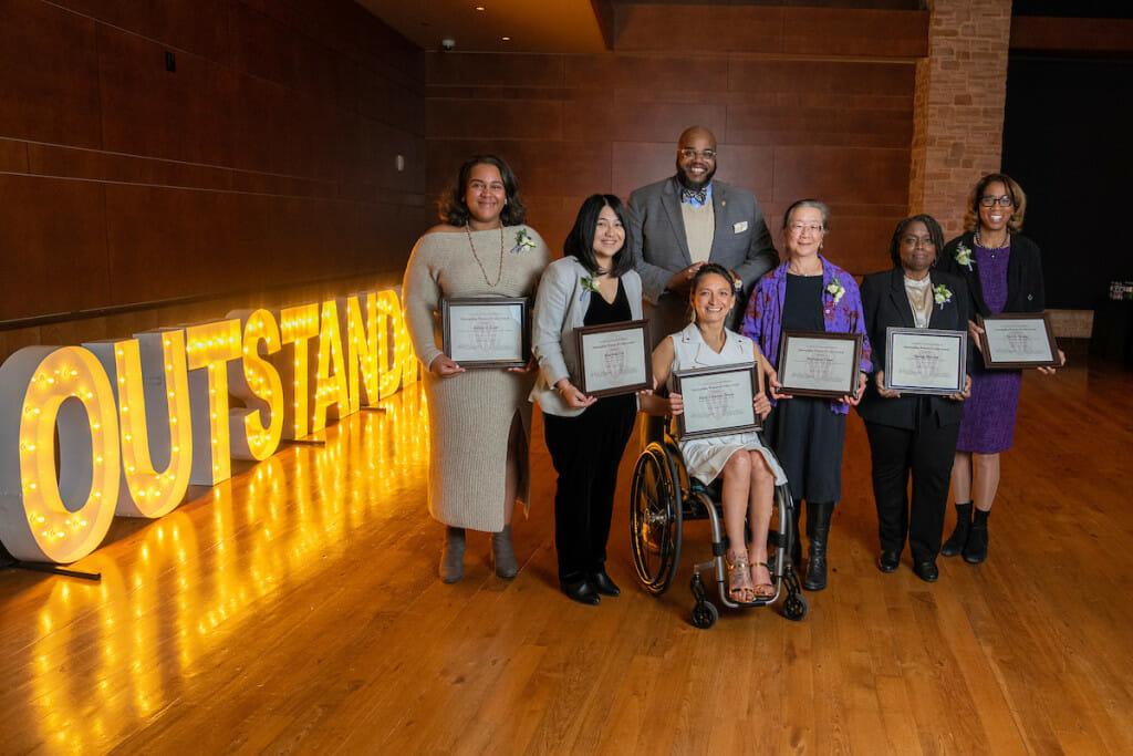 Six woman stand on a stage with a lit-up "Outstanding" sign next to them. A man stands behind them.