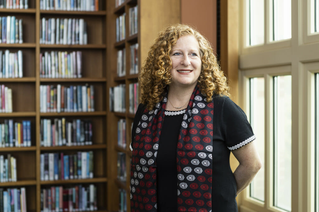 UW–Madison Chancellor Jennifer L. Mnooking stands in the chancellor's suite in Bascom Hall. She is wearing a black dress with a black, red and white scarf adorned with a repeating pattern of UW crests. She is smiling and looking off camera to a sunlit window.