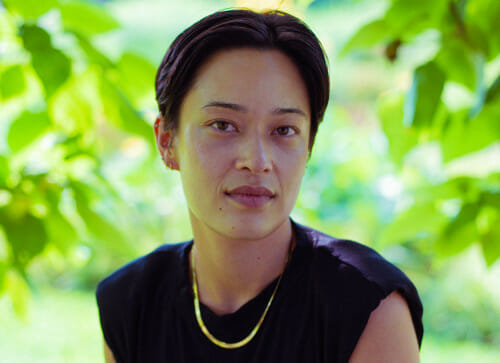 A headshot portrait of Sabrina Imbler. They have short black hair and brown eyes and are looking to the camera with a neutral expression. They are wearing a black sleeveless shirt and a gold necklace.