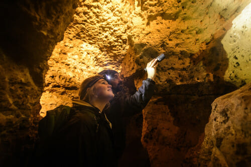 A man and woman stand in a well-lit cavern. The man holds a small flashlight and points to something above their heads.