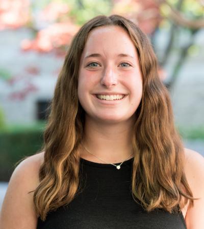 Headshot of Mary Solokas, shot outdoors against a blurred foliage background. Solokas has long brown hair and wears a black sleeveless blouse. She is smiling toward the camera.