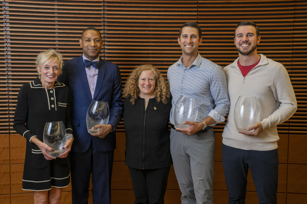 Five people stand looking at the camera, four holding trophies.