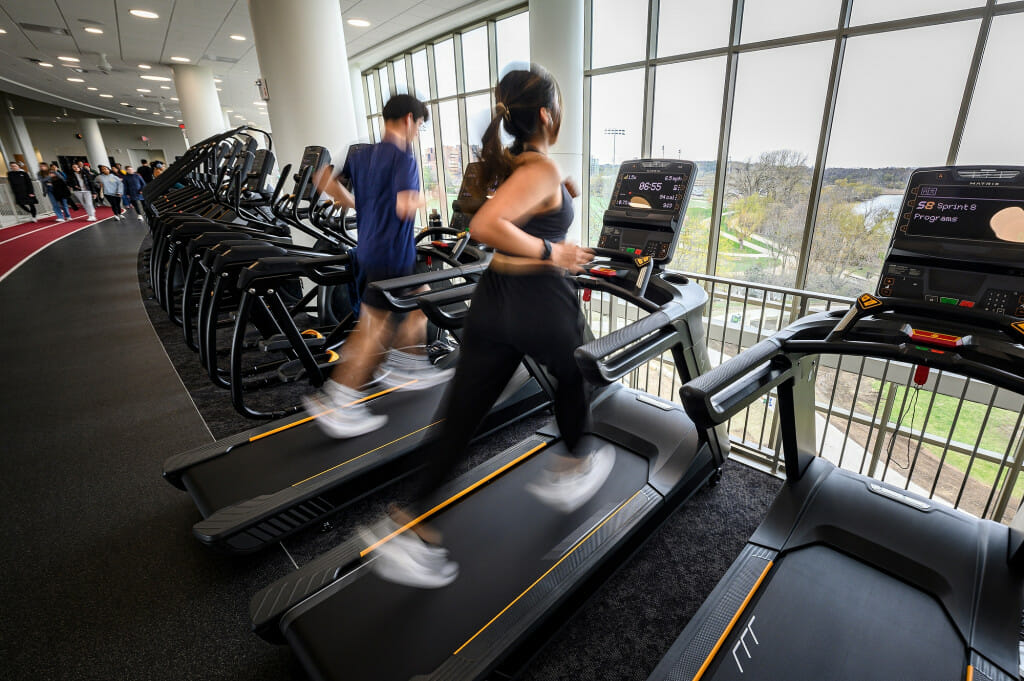 Several people run on treadmills as they look at a lake view out a large window.