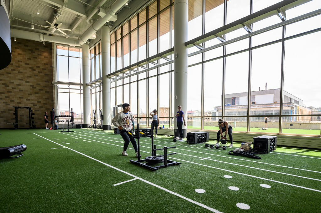 A man sits on a piece of exercise equipment with big windows in the background.