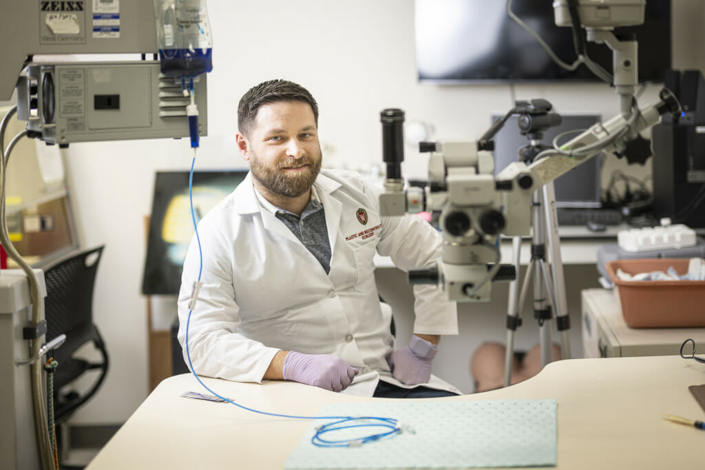 A man in a white lab coat sits at a desk near equipment.