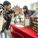 Two men lean over a table to look at a display. A third man stands behind the display as he speaks to the visitors at his table. They are outdoors on Library Mall on a chilly spring day.