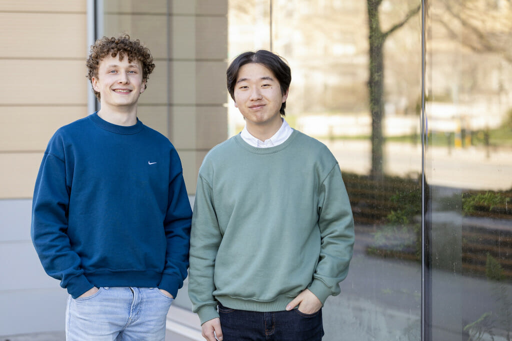 Carl Shirley (left) and Paul Chung (right) stand outside a large plate glass window reflecting the street in front of them.