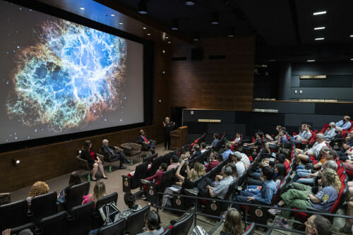 A lecture hall full of people look at a picture of the universe on the big screen.