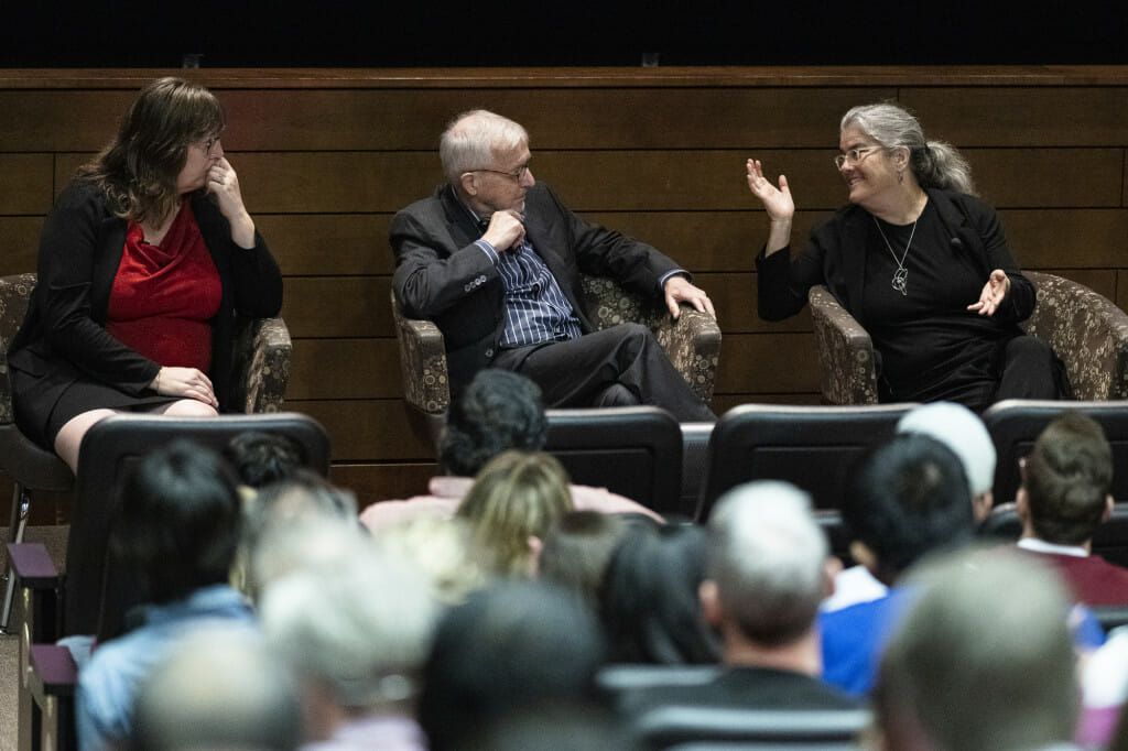 Three people sitting in chairs on stage gesticulate as they talk.