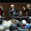 Three people sitting in chairs on stage gesticulate as they talk.