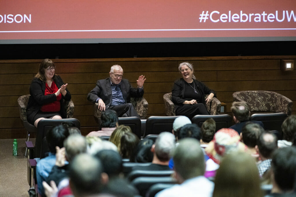 Three people sitting on chairs on stage gesticulate as they talk.