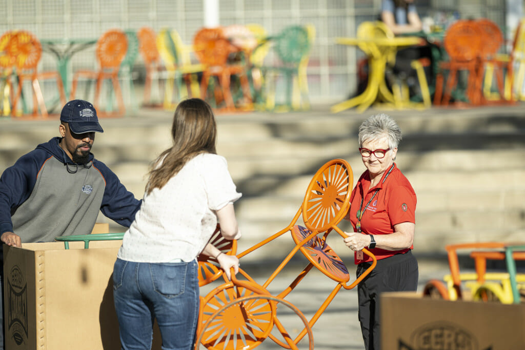 Three people lift chairs from a trolly to place them at tables on the Memorial Union Terrace.