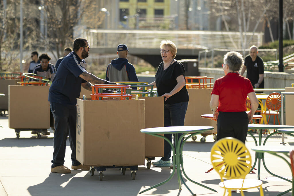 A member of the chair delivery team and Dawn Dever smile and talk as they push trollies of metal chairs across the Memorial Union Terrace.