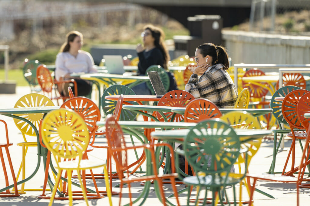 Student Emily Chin wears Air Pods and works on her laptop at a table on the Memorial Union Terrace on a sunny day.