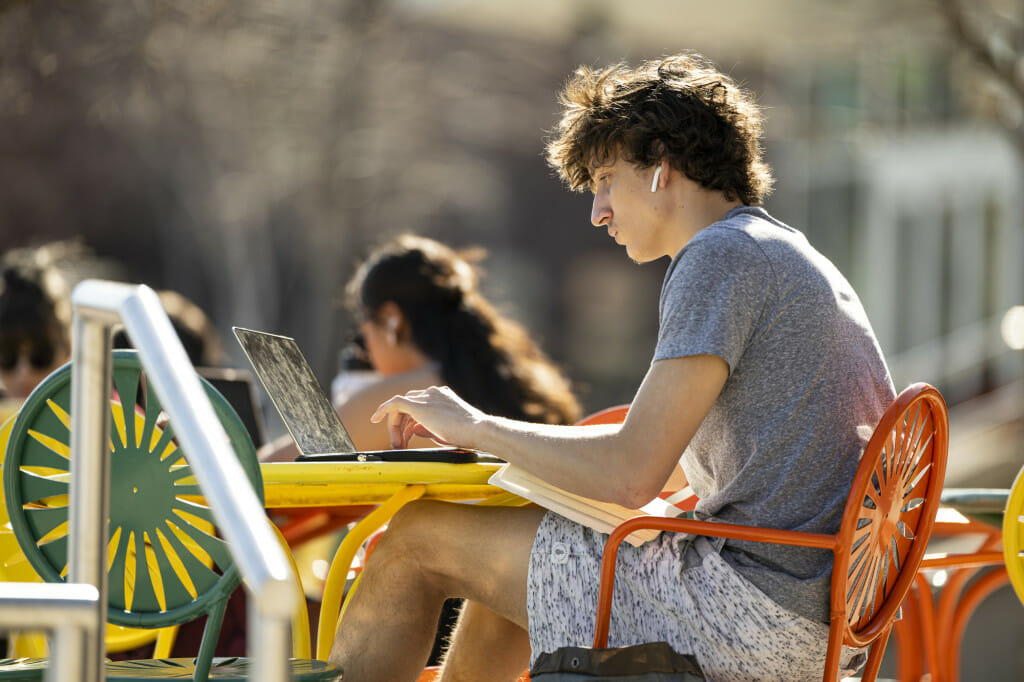 Student Quincy Fields works wears Air Pods and holds a book open on his lap while he works on his laptop, sitting at a table on the Memorial Union Terrace on a sunny day.