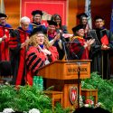 Jennifer Mnookin stands on stage with a crowd of dignitaries, all wearing academic regalia. Mnookin folds her hands on her heart in a gesture of gratitude as she makes an address from a wooden podium with the word 