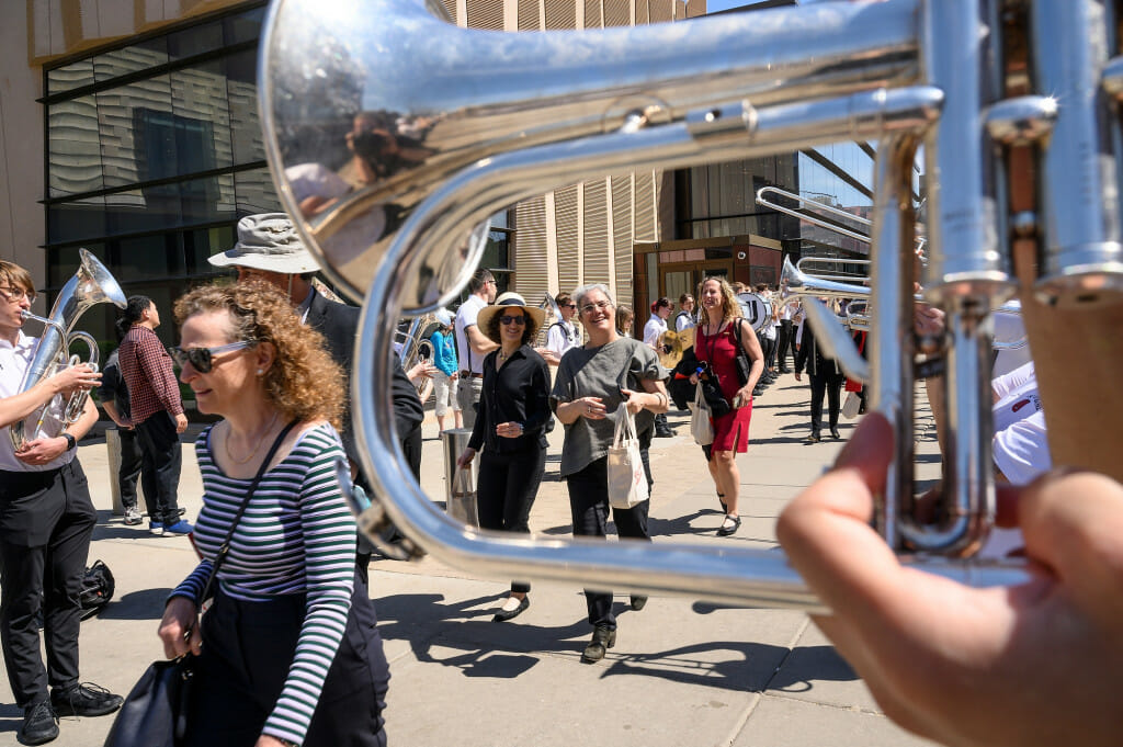 Seen through the curve of a fluglehorn, celebrants walk down a sidewalk with band members playing instruments on either side of them.