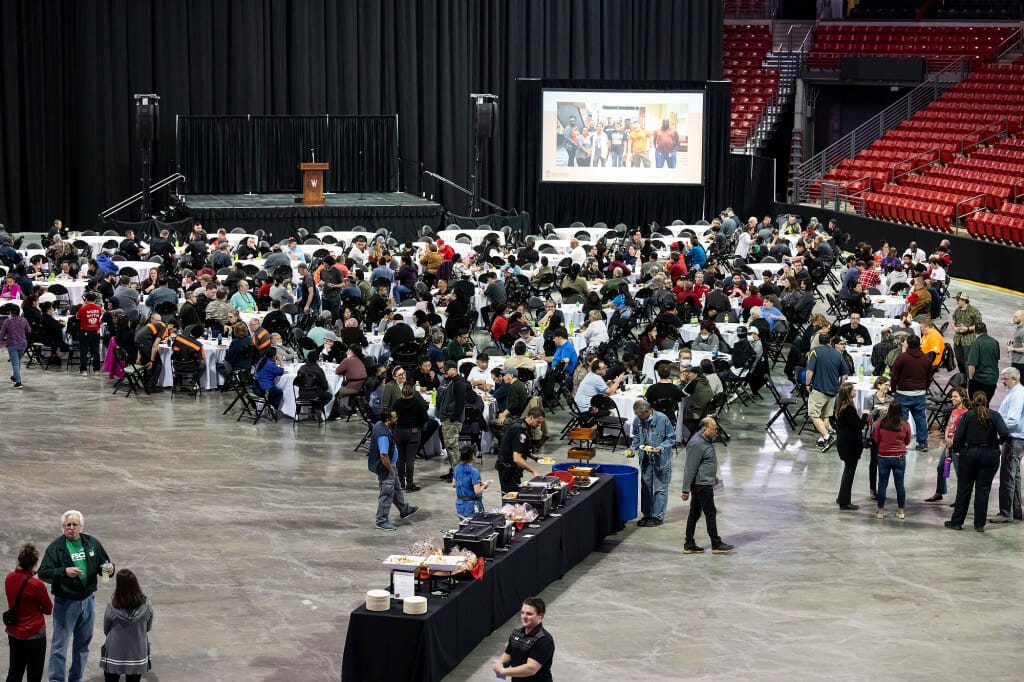 A wide shot of the floor of the Kohl Center arena set up for a banquet reception. There are large tables with chairs, a buffet line and a stage with a podium.