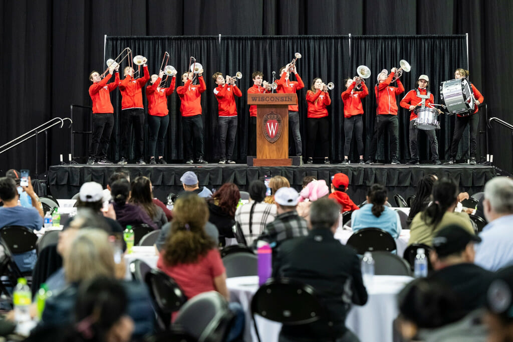 Members of the UW Band's brass section and drum line perform on stage in front of a seated crowd in the Kohl Center arena.