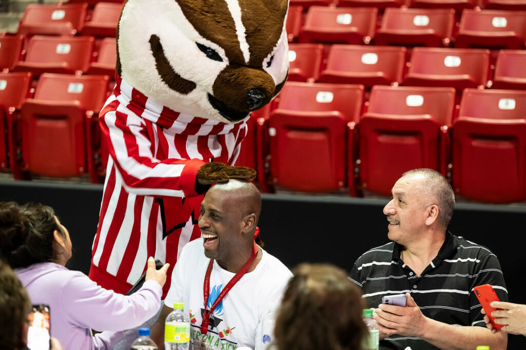 Bucky Badger goofs around with staff attending a second- and third-shift recognition event at the Kohl Center, patting the head of Troymando Pace while colleagues laugh and look on.