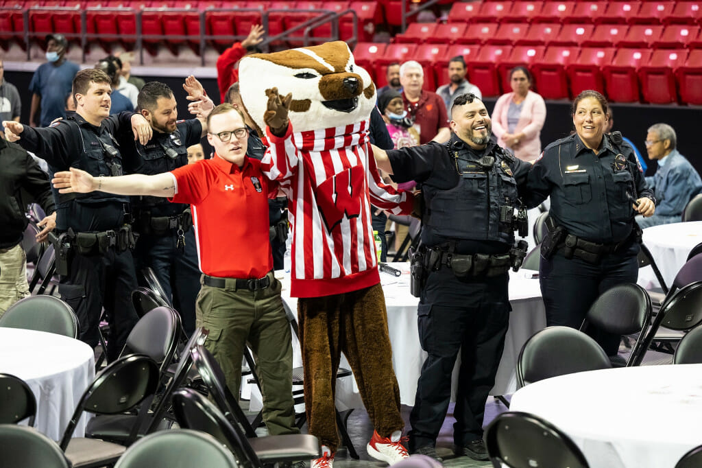 On the floor of the floor of the Kohl Center arena, Bucky Badger and second- and third- shift staff, including UWPD officers in uniform, stand with arms around each other's shoulders as they smile and sing 