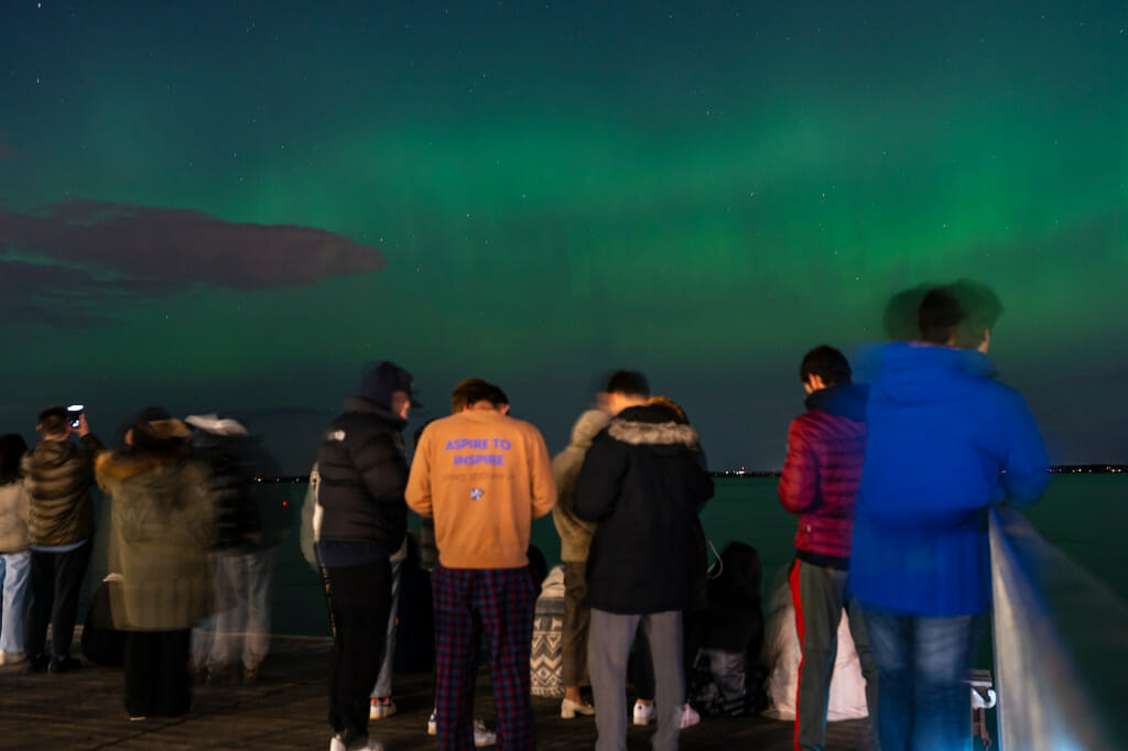 A half dozen students stand on a pier and look at green, wavy lights in the sky.