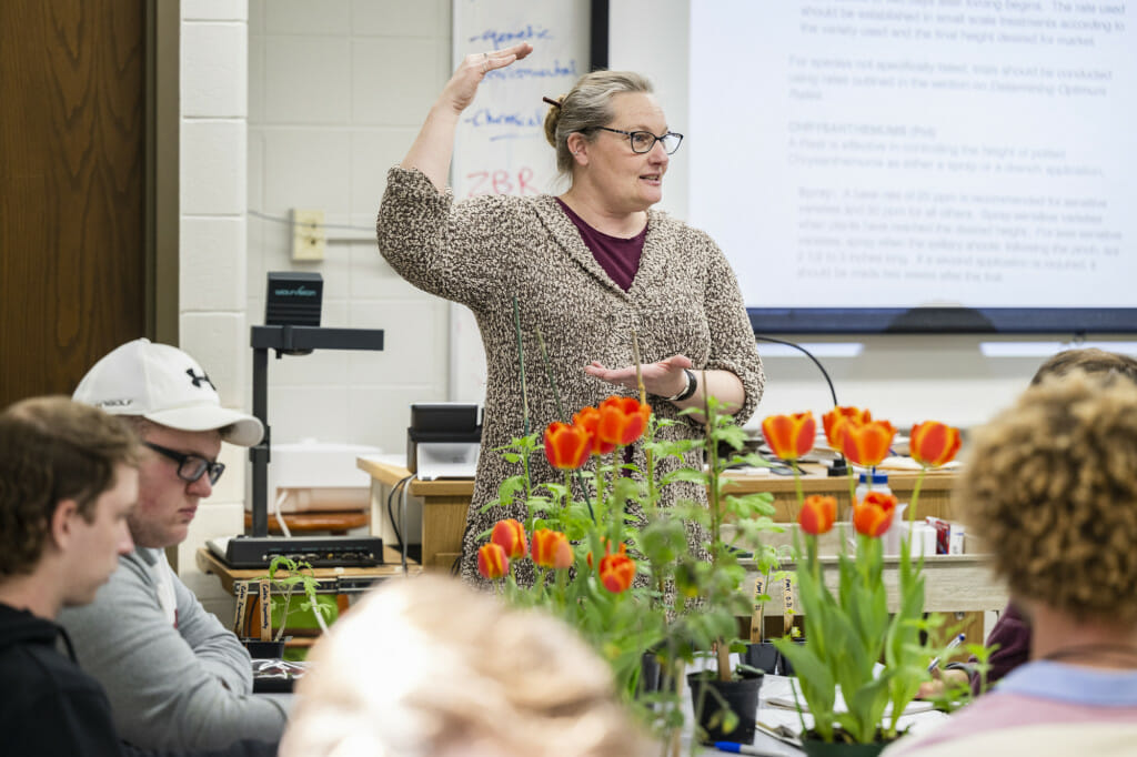 A woman gesticulates as she speaks in front of a whiteboard, with some plants in the foreground.