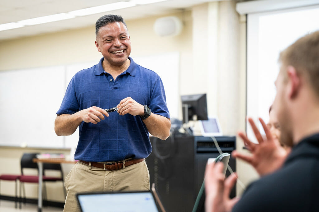 A man, standing, smiles as he talks to a seated student.