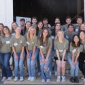 A group of college students stand in three rows outside of a farm outbuilding. They are wearing matching green T-shirts and jeans.