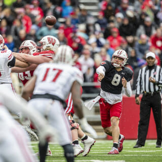 A man in a red jersey flings a football over men in white football uniforms.