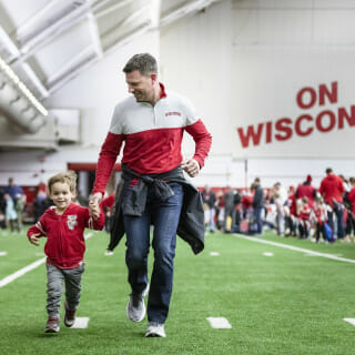 A man and young boy hold hands as they run; an "On Wisconsin" sign is visible in the background.