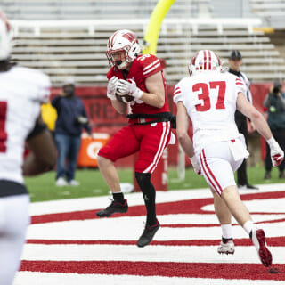 A player with a red jersey cliutches the football to his chest as two players in white jerseys run toward him.