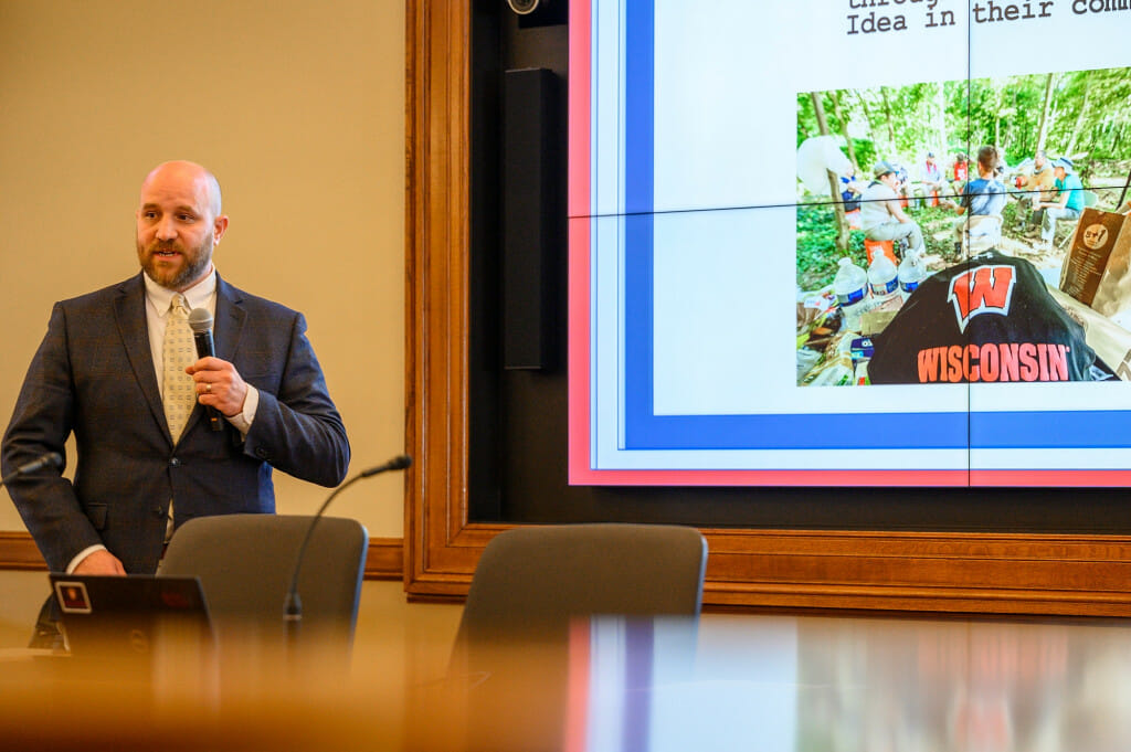 A man standing in front of a conference table and before a large projection screen speaks to an audience.