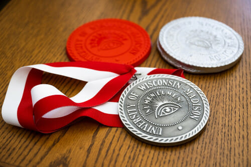 The recast pewter medallion sits on a table alongside a red 3d-printed model and another version cast in aluminum.