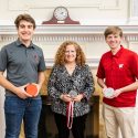 Mechanical Engineering undergraduate students Teekay Kowalewski (left), Jennifer Mnookin (center) and Dylan Zinkgraf (right) face the camera, standing in front of a white, neoclassical fireplace with a clock on the mantle. Kowalewski holds a 3D-printed version of the medallion. Mnookin holds the new, pewter medallion. Zinkgraf holds an aluminum rendition of the medallion.